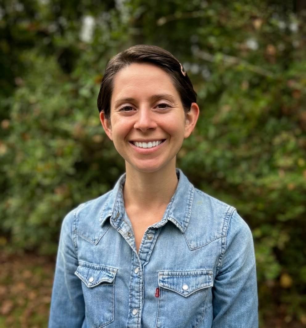 Portrait of Stacey Fronek smiling outdoors in a denim shirt, with soft green foliage in the background